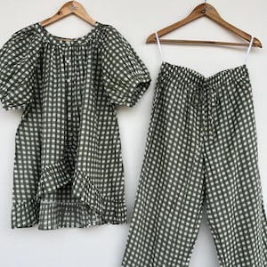 May include: A two-piece set featuring a button-down blouse with puff sleeves and a ruffled hem, paired with matching wide-leg trousers that have a drawstring waist and ruffled cuffs. Both pieces are made of olive green and white gingham fabric.