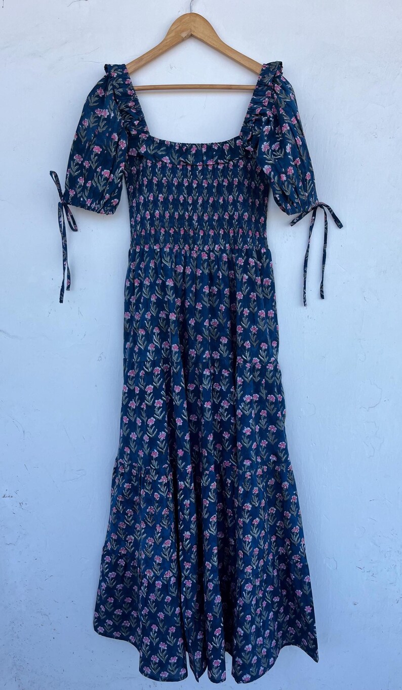 May include: A navy blue floral print dress with a square neckline and short sleeves. The dress features a smocked bodice and a flowing skirt. The sleeves are tied with matching fabric ties. The dress is adorned with a repeating pattern of small pink flowers.