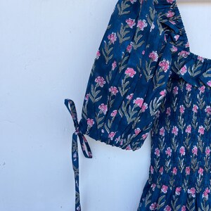 May include: A navy blue dress with pink floral print. The dress features puffed sleeves with tie details and a square neckline. The fabric has a textured appearance, and the floral pattern includes green leaves.