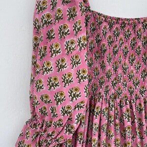May include: Pink dress with a floral pattern featuring yellow flowers and green leaves. The dress has a square neckline and puffed sleeves with ruffled cuffs. The bodice is smocked, and the fabric is a soft, lightweight material.