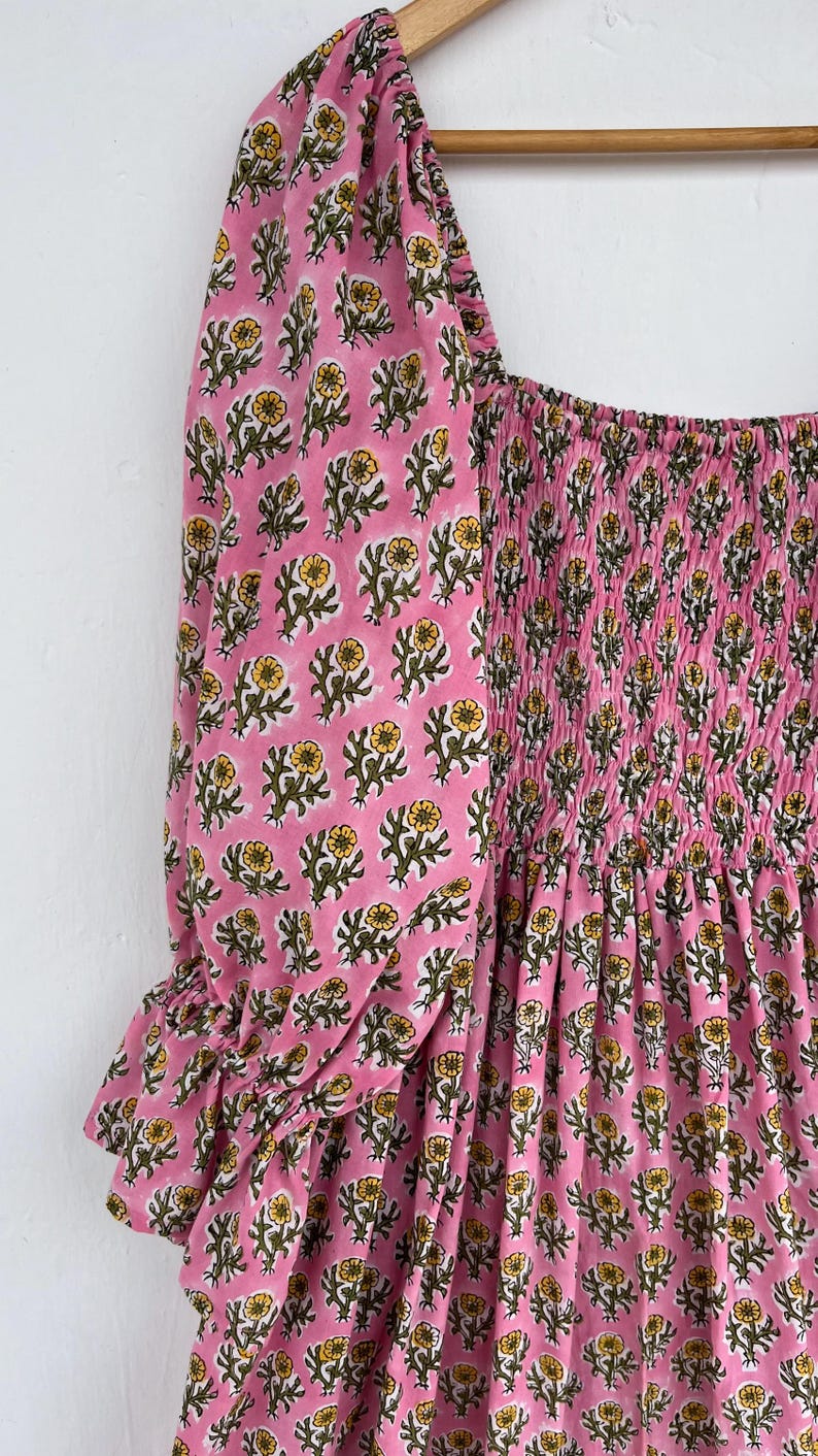 May include: Pink dress with a floral pattern featuring yellow flowers and green leaves. The dress has a square neckline and puffed sleeves with ruffled cuffs. The bodice is smocked, and the fabric is a soft, lightweight material.