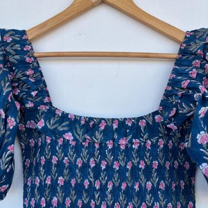 May include: A dark teal dress with a floral pattern of pink flowers and green leaves. The dress has a square neckline and short, puffed sleeves. The fabric is gathered at the bodice and shoulders, and is hanging on a wooden hanger.