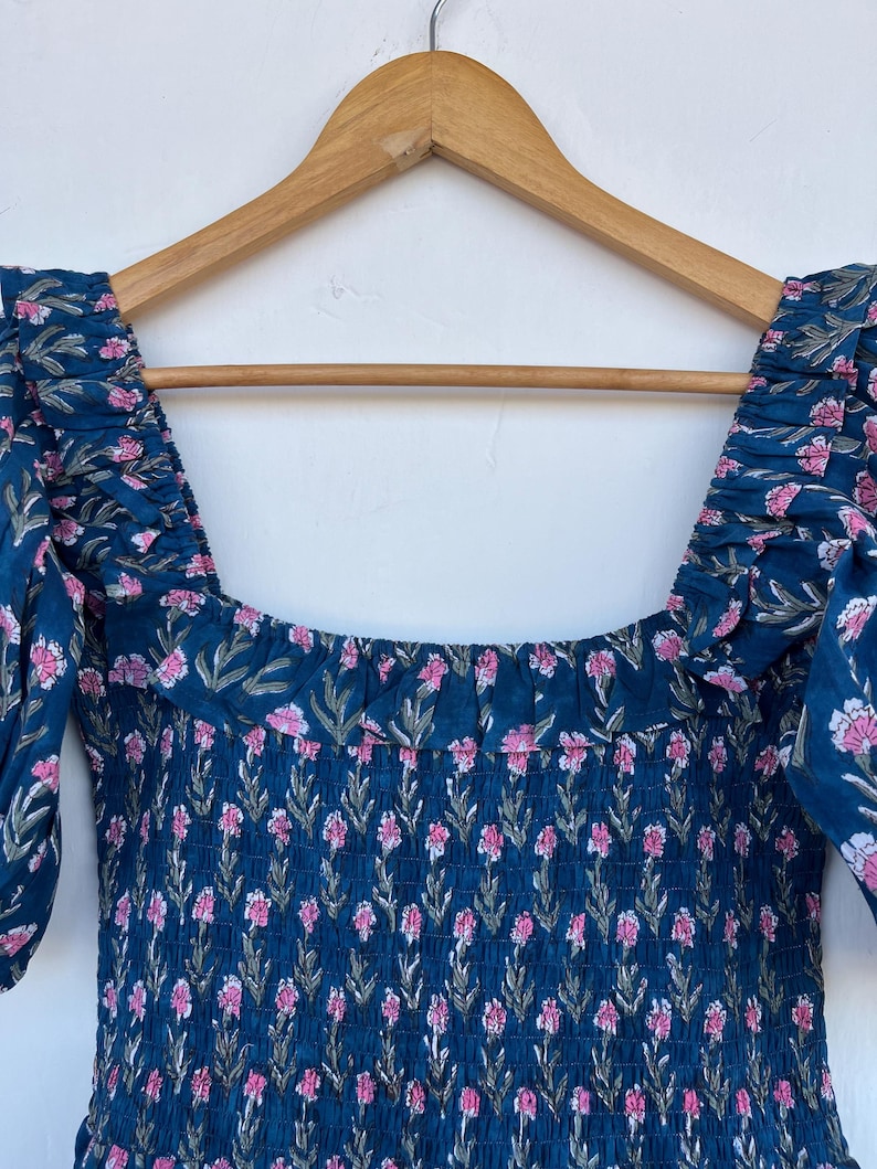 May include: A dark teal dress with a floral pattern of pink flowers and green leaves. The dress has a square neckline and short, puffed sleeves. The fabric is gathered at the bodice and shoulders, and is hanging on a wooden hanger.