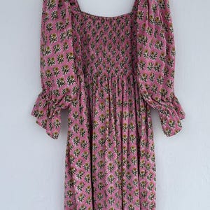 May include: A pink floral print dress with a square neckline and three-quarter sleeves. The dress features a smocked bodice and a flowing skirt. The sleeves have a ruffled cuff. The floral pattern includes yellow and green accents.