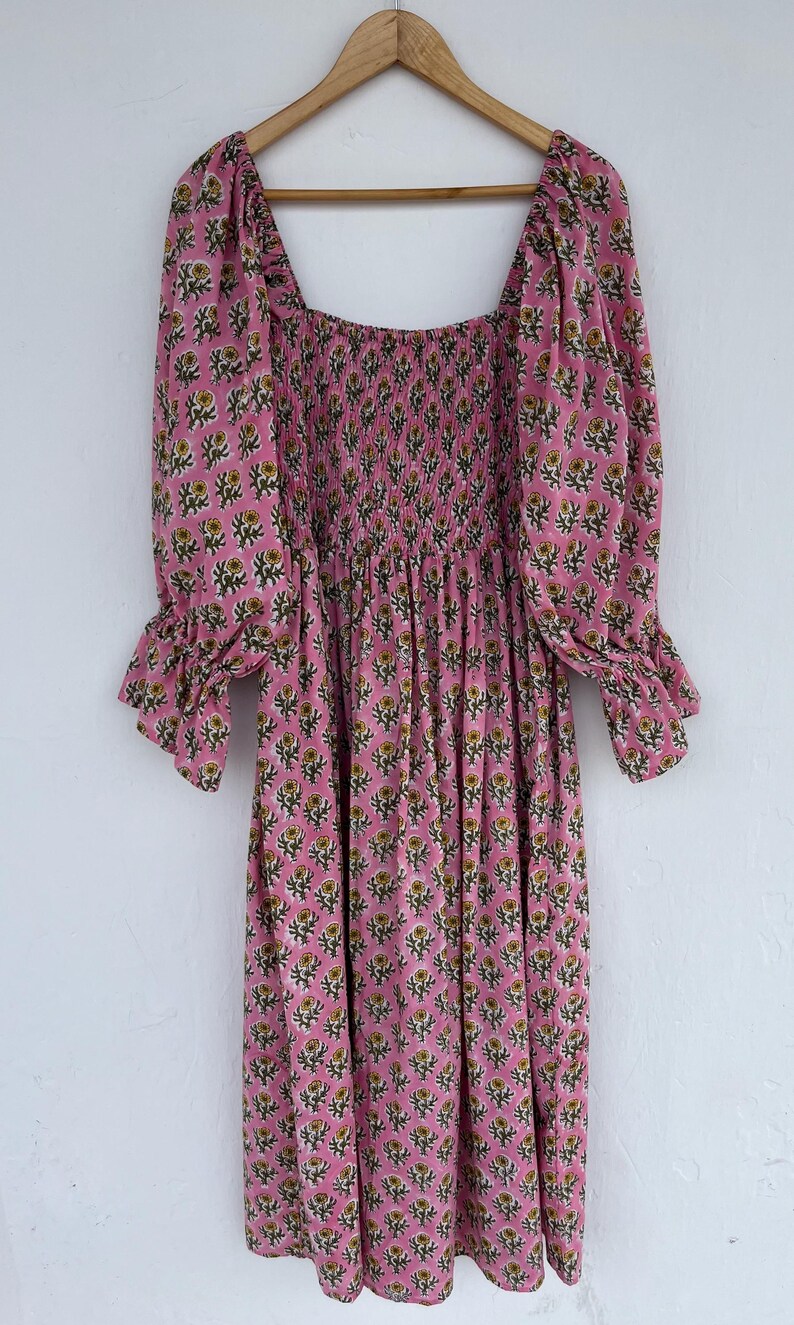 May include: A pink floral print dress with a square neckline and three-quarter sleeves. The dress features a smocked bodice and a flowing skirt. The sleeves have a ruffled cuff. The floral pattern includes yellow and green accents.