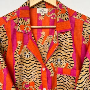 May include: A button-down pajama top with a vibrant tiger print. The shirt features a bold pattern of orange and pink stripes with tiger illustrations. The collar and pocket are trimmed in orange. The shirt is hanging on a wooden hanger.