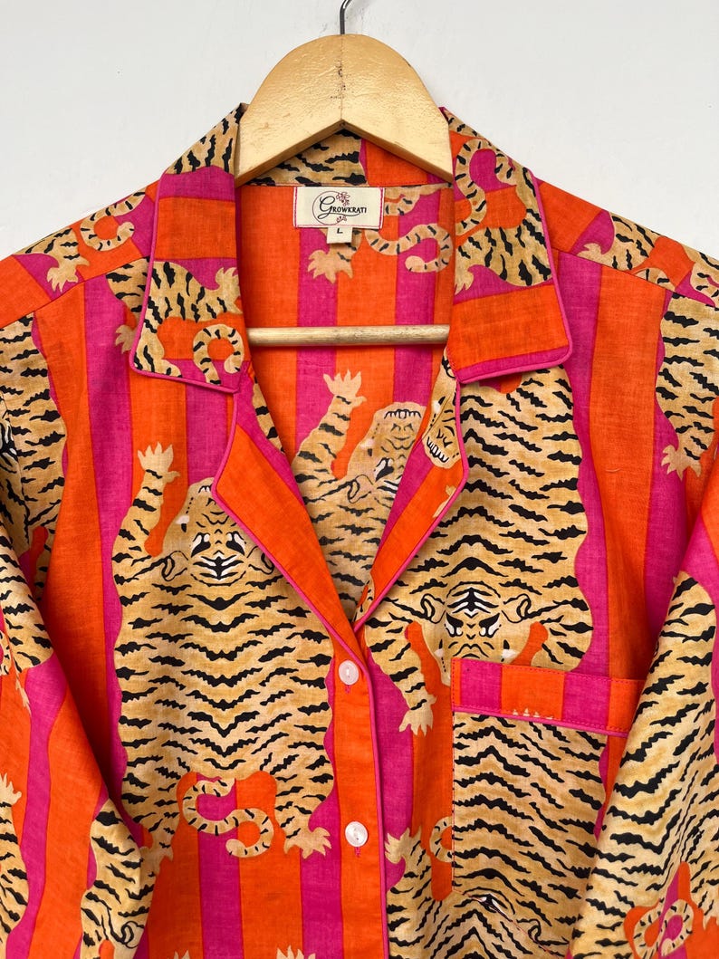 May include: A button-down pajama top with a vibrant tiger print. The shirt features a bold pattern of orange and pink stripes with tiger illustrations. The collar and pocket are trimmed in orange. The shirt is hanging on a wooden hanger.