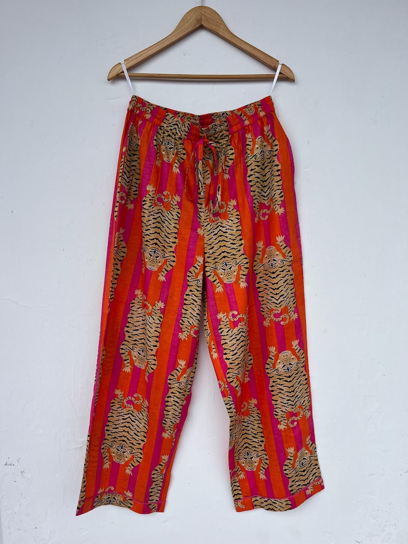 May include: Orange and pink striped pants with a tiger print. The pants are hanging on a wooden hanger. The pants have a drawstring waist and a loose, comfortable fit. The tiger print is in shades of brown and black.