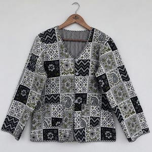 May include: A long-sleeved quilted jacket featuring a patchwork design of black, white, and olive green floral, paisley, and chevron patterns. The jacket has a V-neckline, a front button closure with four white buttons, and two square front pockets.