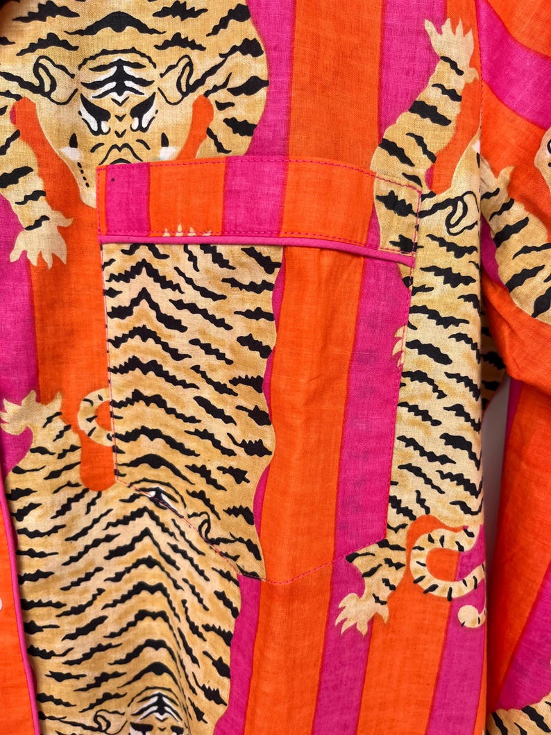 May include: Close-up of a vibrant pajama top featuring a tiger print and vertical stripes in orange, pink, and fuchsia. The pocket is outlined in pink. The tiger print includes black stripes on a tan background.