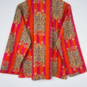May include: An orange and pink striped button-down shirt with a repeating tiger print. The long-sleeved shirt is hanging on a wooden hanger against a white background. The tiger print features a tan tiger with black stripes.