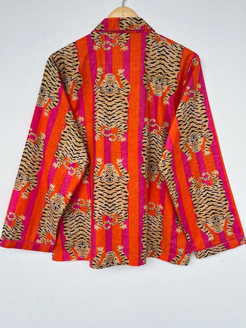 May include: An orange and pink striped button-down shirt with a repeating tiger print. The long-sleeved shirt is hanging on a wooden hanger against a white background. The tiger print features a tan tiger with black stripes.