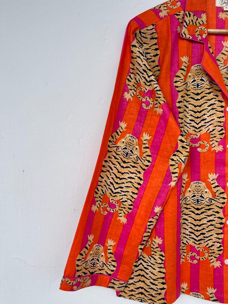May include: Orange and pink striped pajama top with a tiger print. The long-sleeved shirt features a collared neckline and button-down front. The tiger print is in a tan and black color.