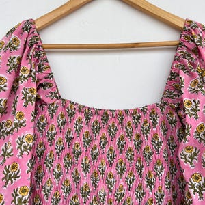 May include: A pink dress with a square neckline and puffed sleeves, adorned with a floral pattern in yellow and green. The bodice features a smocked design. The dress is hanging on a wooden hanger.