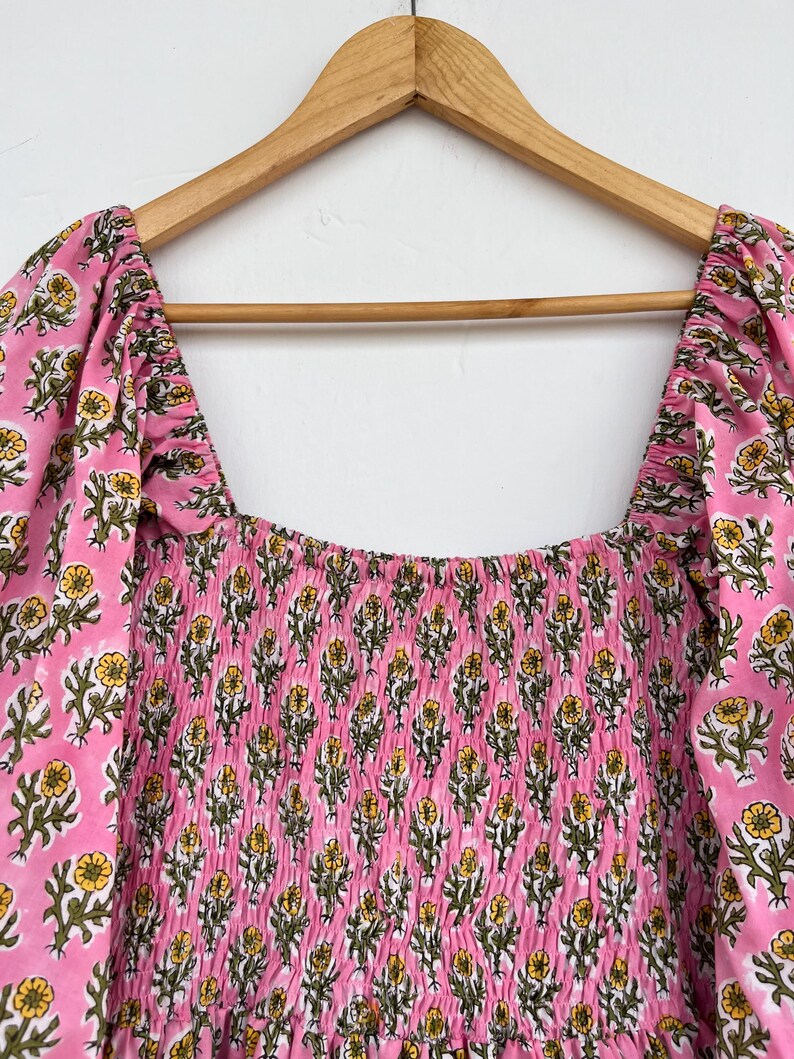 May include: A pink dress with a square neckline and puffed sleeves, adorned with a floral pattern in yellow and green. The bodice features a smocked design. The dress is hanging on a wooden hanger.