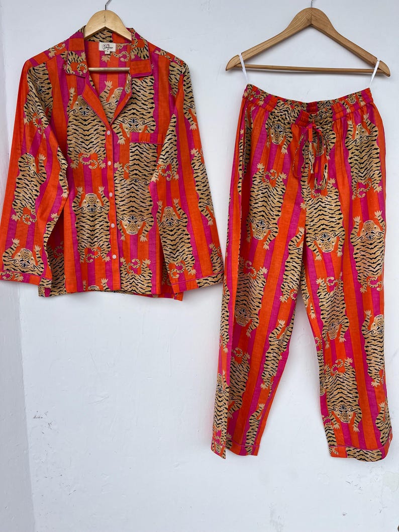 May include: Orange and pink striped pajama set with a tiger print. The set includes a button-down shirt and drawstring pants. The shirt has a collar and a breast pocket. The pants have a drawstring waist. The pajamas are hanging on a wooden hanger.