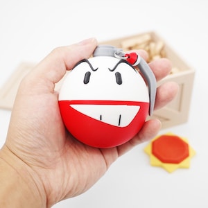 Puede incluir: Un llavero redondo de novedad con un diseño de bomba de dibujos animados. La mitad superior es blanca con ojos y cejas negras, y la mitad inferior es roja. Tiene un anillo de tracción gris y un acento rojo.