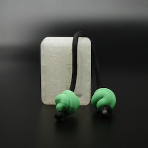May include: A clear, rectangular case with the word "Metamorphosis" etched on the front. A black cord is attached, with two green, spiral-shaped objects at the ends. The objects are on a black background.