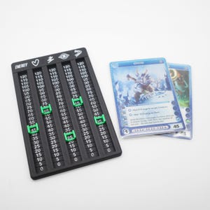 May include: A black plastic game tracker with five vertical columns labeled with icons for energy, heart, lightning, eye, and arrow symbols. Each column features numbers from 0 to 120 with movable green sliders. Two blue trading cards are placed beside it.