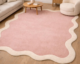 May include: A large, pink rug with a cream-colored, wavy border. The rug is placed on a hardwood floor in a living room setting. The room also features a sofa, armchair, and wooden furniture.
