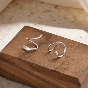 May include: A pair of silver-toned earrings displayed on a small, dark wooden block. One earring has a spiral design, while the other is a curved shape. The earrings are set against a neutral background, with soft lighting.