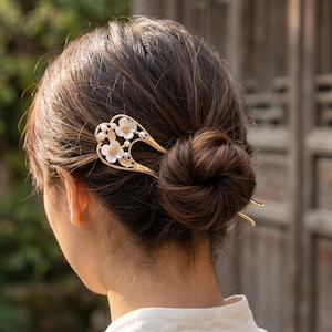 Puede incluir: Una horquilla dorada con diseño floral, con flores blancas y detalles de perlas. El accesorio para el cabello está insertado en un moño, mostrando el elemento decorativo. El diseño de la horquilla incluye un elemento en forma de corazón.