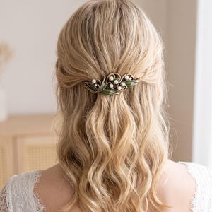 Peut inclure: Une barrette à cheveux dotée d'une monture dorée aux motifs tourbillonnants complexes, agrémentée de petites perles blanches sphériques et de détails en émail vert en forme de feuilles, fixée dans des cheveux blonds ondulés.