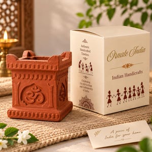 May include: A square terracotta diya holder featuring an embossed Om symbol on the front, accompanied by a cream-colored cardboard box labeled Ornate India Indian Handicrafts with tribal illustrations and a small card reading A piece of India for your home.