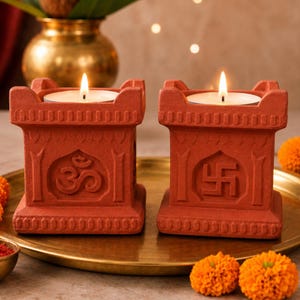 May include: Two terracotta-coloured clay candle holders with square bases and crenelated tops. Each holder features a central embossed symbol, one displaying an Om and the other a swastika. A lit white tea light candle sits inside each holder.