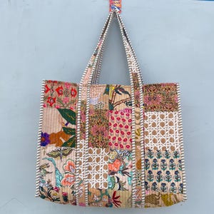Puede incluir: Un bolso tote acolchado con un diseño de patchwork de varios patrones florales, botánicos y geométricos en tonos beige, rosa, verde y azul. El bolso tiene dos asas largas para el hombro con un borde a rayas blancas y beige en los extremos.