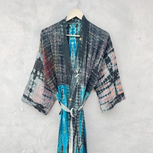 May include: A tie-dye robe in shades of black, grey, blue, and red hangs on a wooden hanger. The robe features a wrap-around design with a matching belt. The fabric has a patchwork appearance with embroidered details.