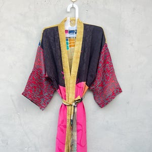 May include: A long-sleeved robe featuring a patchwork design with a black upper section, bright pink lower panels, and red patterned sleeves. The garment includes a mustard yellow trim along the front opening and a matching tie belt at the waist.