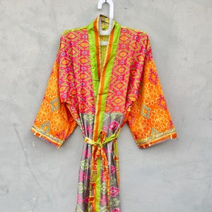 May include: A patterned kimono robe with a tie belt. The robe features orange, pink, green, and grey geometric designs. The robe is hanging on a white hanger against a grey background.