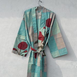 May include: A patterned robe in shades of teal, brown, and red hangs against a gray wall. The robe features a checkered pattern with floral accents and a matching belt. The robe is made of a lightweight fabric.