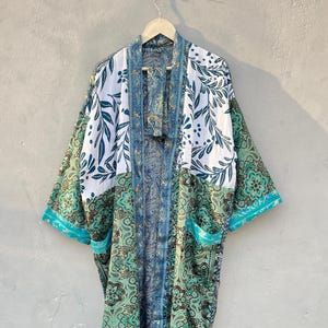 May include: A long-sleeved kimono robe featuring a patchwork design with a white upper section adorned with dark teal leaf patterns and a lower section displaying a complex green, blue, and black paisley and floral print with a matching belt.