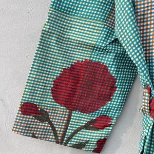 May include: A teal and orange checkered robe sleeve with a large red floral design. The floral design includes a large flower and several buds. The robe is made of a lightweight fabric.