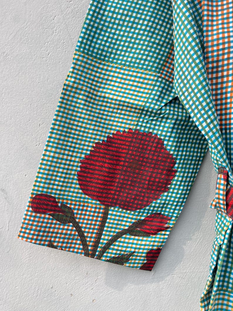 May include: A teal and orange checkered robe sleeve with a large red floral design. The floral design includes a large flower and several buds. The robe is made of a lightweight fabric.