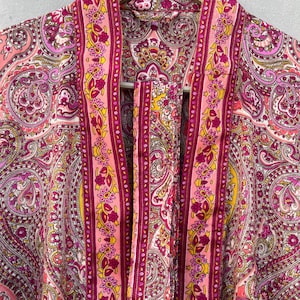Puede incluir: Bata rosa con estampado de cachemira y corbata a juego. La bata presenta un dise&ntilde;o vibrante con tonos de rosa, morado y amarillo. La bata est&aacute; colgada de una percha de madera.