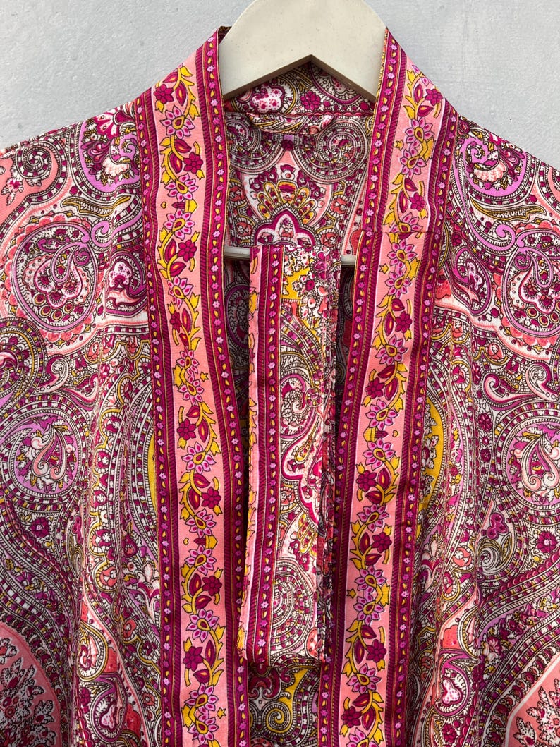 Puede incluir: Bata rosa con estampado de cachemira y corbata a juego. La bata presenta un dise&ntilde;o vibrante con tonos de rosa, morado y amarillo. La bata est&aacute; colgada de una percha de madera.