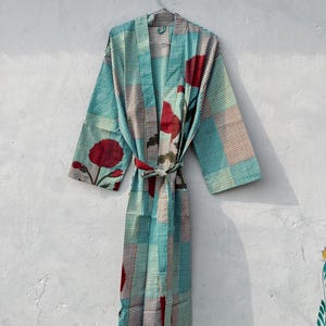 May include: A patterned robe with a tie waist, featuring a teal and brown checkered design with large red floral accents. The robe is hanging on a metal hanger against a light gray wall. The robe's sleeves are wide and the fabric appears lightweight.