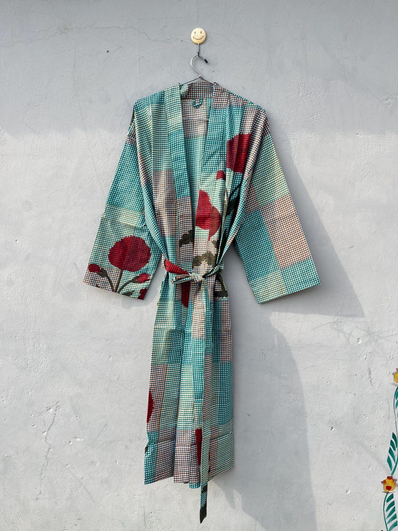 May include: A patterned robe with a tie waist, featuring a teal and brown checkered design with large red floral accents. The robe is hanging on a metal hanger against a light gray wall. The robe's sleeves are wide and the fabric appears lightweight.
