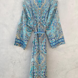 May include: A long, flowing robe with a vibrant paisley pattern in shades of blue, teal, and gold. The robe features long sleeves, a collared neckline, and a matching belt. The garment is hanging on a hanger against a neutral gray background.