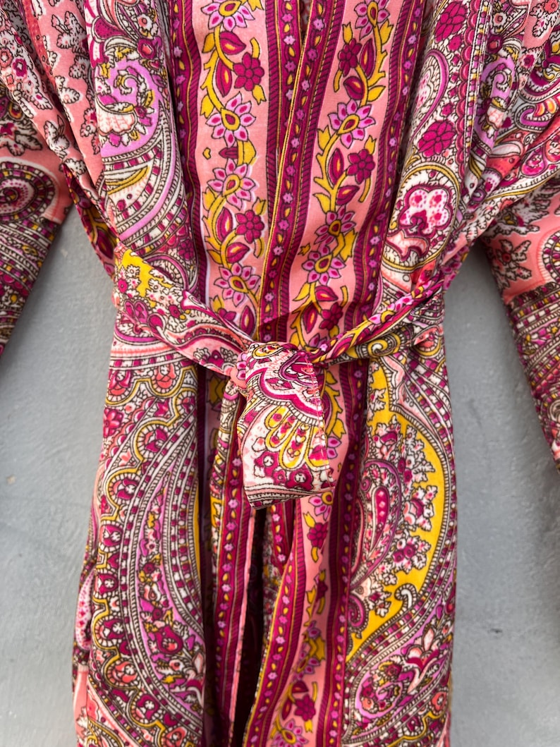 Puede incluir: Una bata con estampado de cachemira en tonos rosa, amarillo y morado. La bata est&aacute; atada a la cintura con un cintur&oacute;n a juego. La tela presenta un dise&ntilde;o floral detallado y rayas verticales. Las mangas son visibles a ambos lados de la bata.
