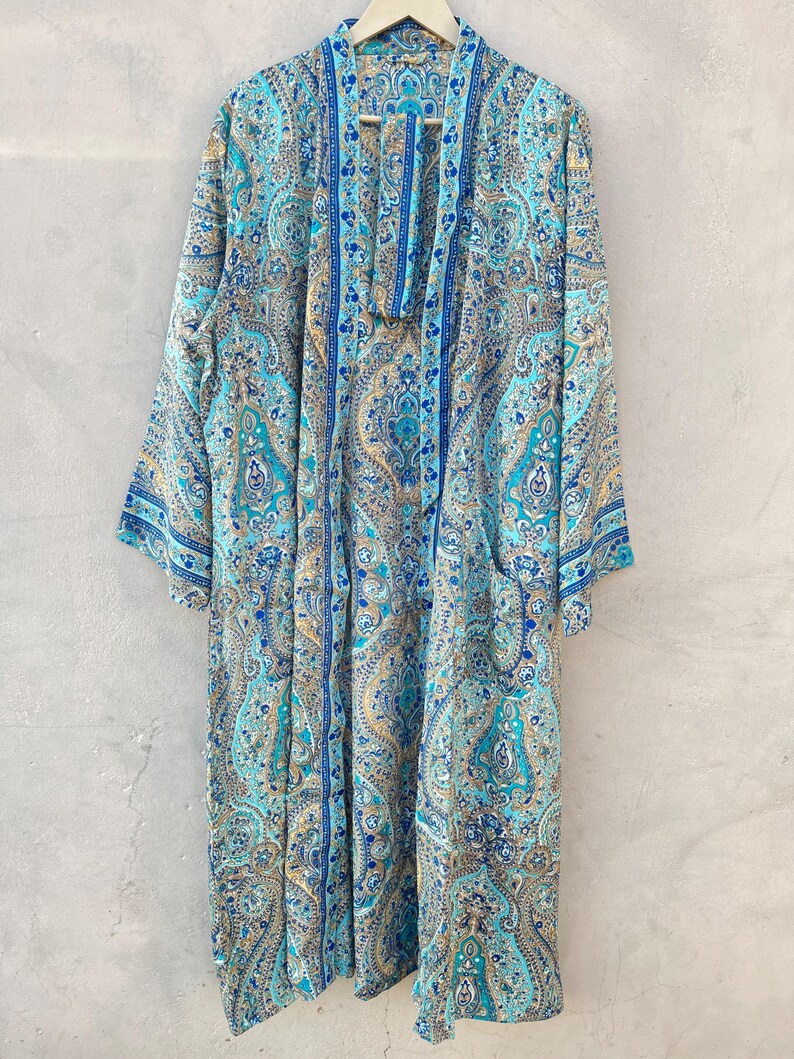 May include: A long, flowing robe with a paisley pattern in shades of blue, teal, and gold. The robe has long sleeves and a draped collar. It is hanging on a wooden hanger against a neutral background.