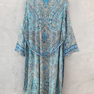 May include: A long, flowing robe with a paisley pattern in shades of blue, teal, and gold. The robe has long sleeves, a collar, and a matching belt. The garment is hanging against a neutral gray background.