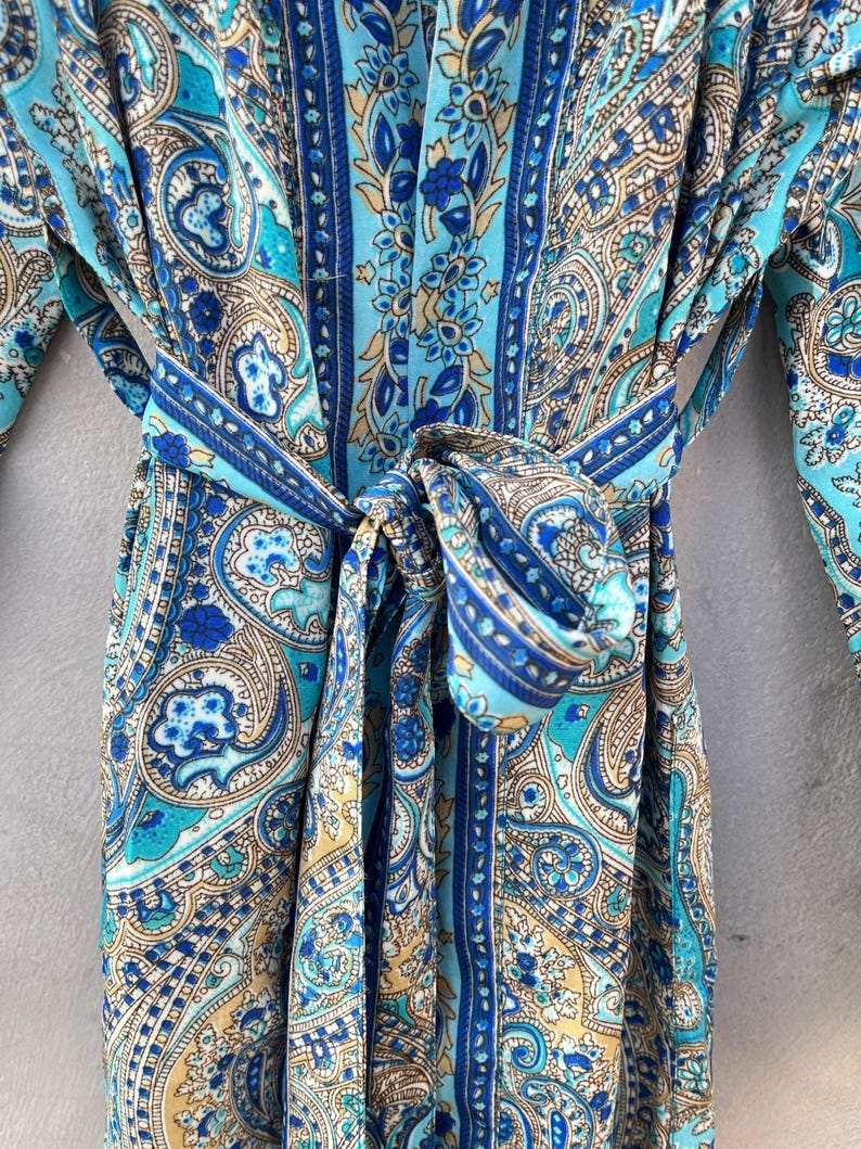 May include: A blue paisley patterned robe with a matching belt tied in a bow. The robe features shades of blue, teal, and tan. The fabric has a silky appearance and a detailed floral design.