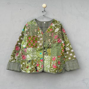 Könnte beinhalten: Eine langärmelige Steppjacke mit Patchwork-Design in Grün-, Rosa- und Weißtönen mit Blumenmustern. Die Jacke hat einen V-Ausschnitt, eine Knopfleiste vorne und breite Ärmelbündchen mit einem schwarz-weißen vertikalen Streifenmuster.