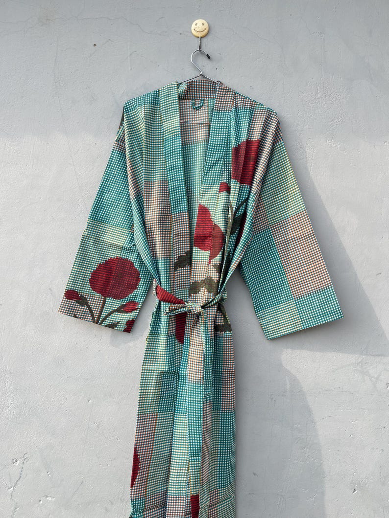 May include: A patterned robe in shades of teal, brown, and red hangs against a gray wall. The robe features a checkered pattern with floral accents and a matching belt. The robe is made of a lightweight fabric.