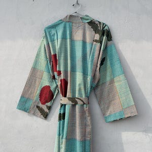 May include: A patterned robe with a belt, hanging on a wall. The robe features a teal, brown, and white checkered pattern with large red and green floral accents. The robe is made of a lightweight fabric.