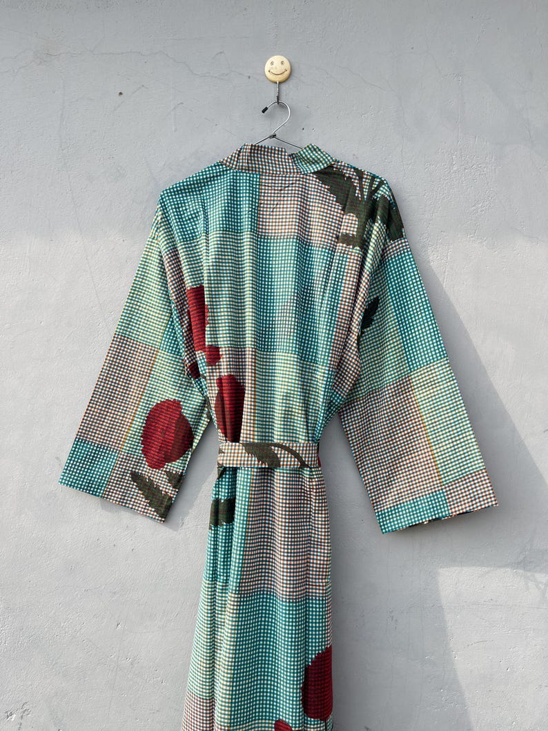 May include: A patterned robe with a belt, hanging on a wall. The robe features a teal, brown, and white checkered pattern with large red and green floral accents. The robe is made of a lightweight fabric.
